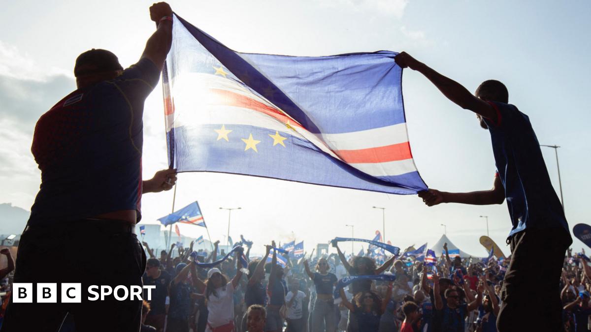 Two men silhouetted hold aloft a Cape Verde flag which is rendered almost translucent as sunlight shines through it as hundreds of people celebrating are seen in front of them waving more flags and holding up scarves