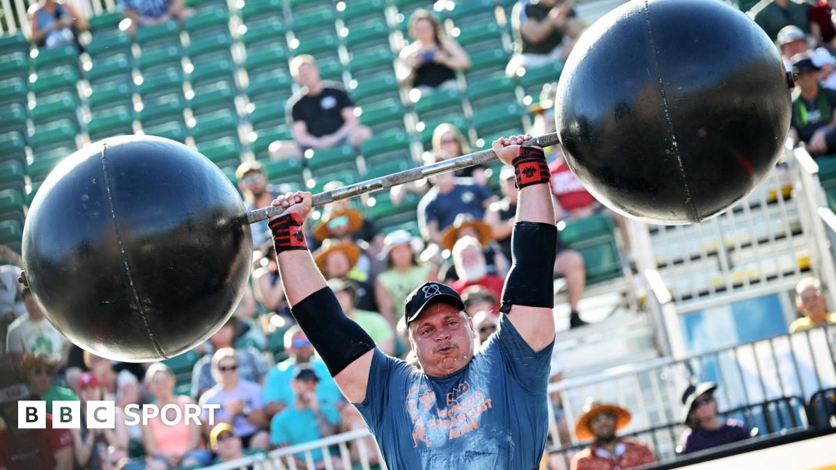 Rayno Nel of South Africa seen from chest up, wearing a blue t-shirt, black cap and black sunglasses, performs the stone to shoulder press at 2025's World Strongest Man in Sacramento. He has a look of concentration and effort on his face as he flexes his huge bicep and raises his right arm to balance an enormous grey stone in the crook of his neck and against his head. Partially seen in the background are two blurred people, one of whom is clapping