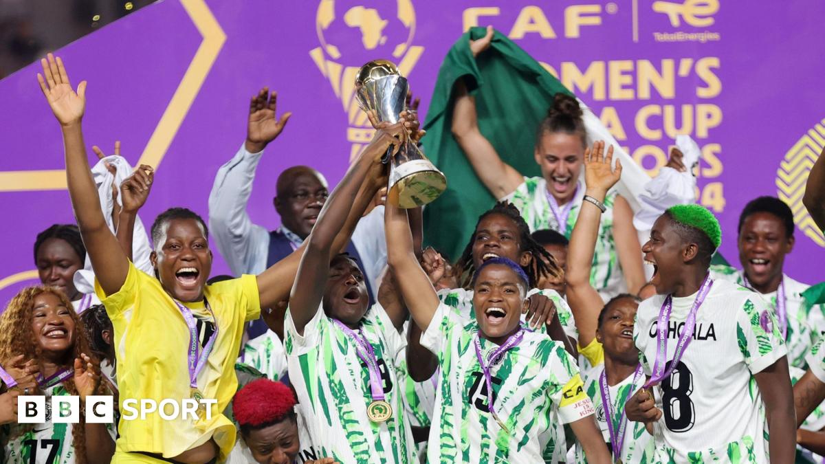 A group of joyous Nigeria players raise their arms and shout for joy as they lift the Women's Africa Cup of Nations trophy during the presentation at the 2024 Wafcon finals in Morocco. The players have golden medals around their necks on purple ribbons, with the trophy being lifted by captain Rasheedat Ajibade, who has hair dyed blue, while goalkeeper Chiamaka Nnodozie, wearing bright yellow kit and raising both arms mid-jump, is one of the players near her skipper