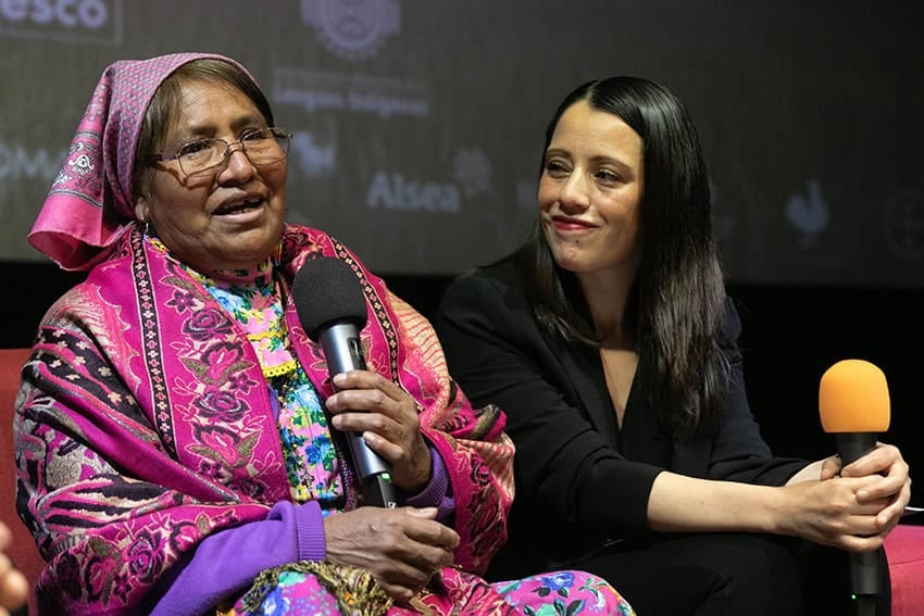 An older woman in colorful Indigenous clothing speaks into a microphone, next to a young Mexican woman dressed in black