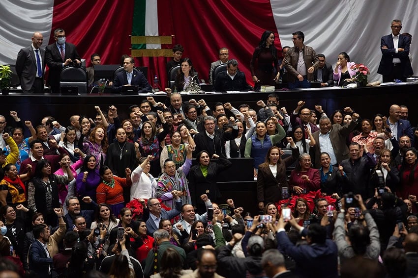Ricardo Monreal stands at a podium in the Mexican Chamber of Deputies (congress chambers) surrounded by dozens of supporters with their fists raised in the air