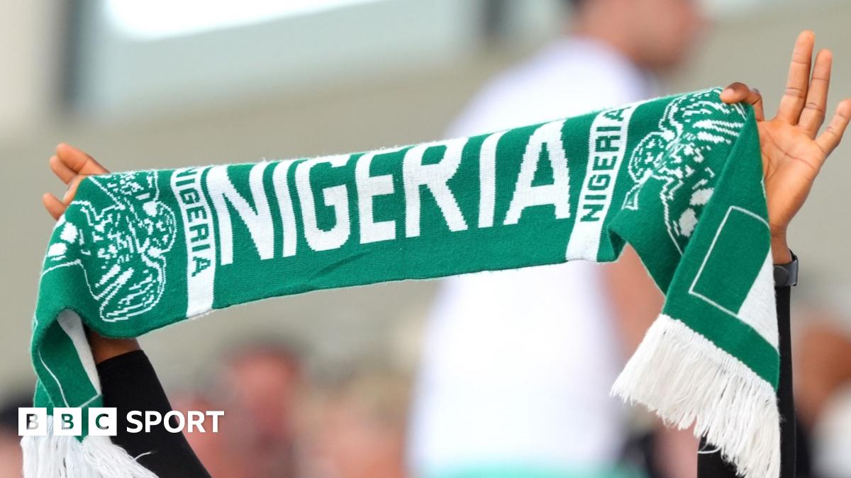 A Nigeria football fan holds a green t-shirt with the words "Hope Dey" (meaning there is hope) written in white, and "we must qualify" underneath. He is surrounded by other Nigeria fans who are seated in a stadium