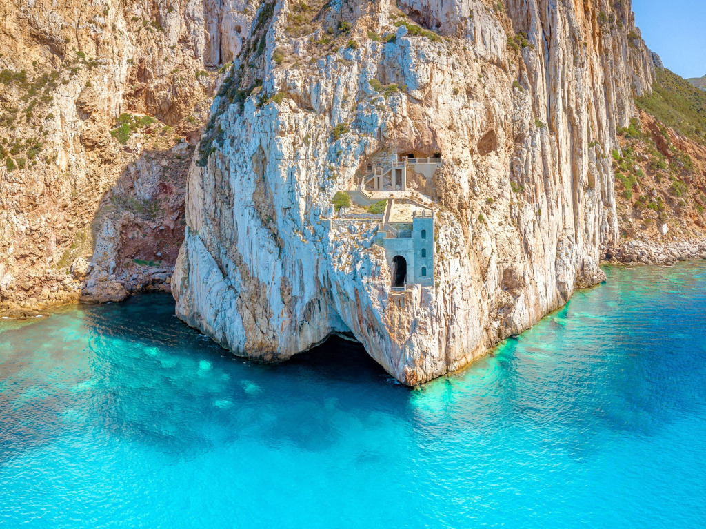 Quick guide to Sardinia. Image shows building built into cliff face at Porto Flavia, Sardinia. Image credit: https://www.goodfon.com/landscapes/wallpaper-cliff-building-porto-flavia-sardinia-italy-biriuzovoe-more-s.html https://creativecommons.org/licenses/by-nc/4.0/