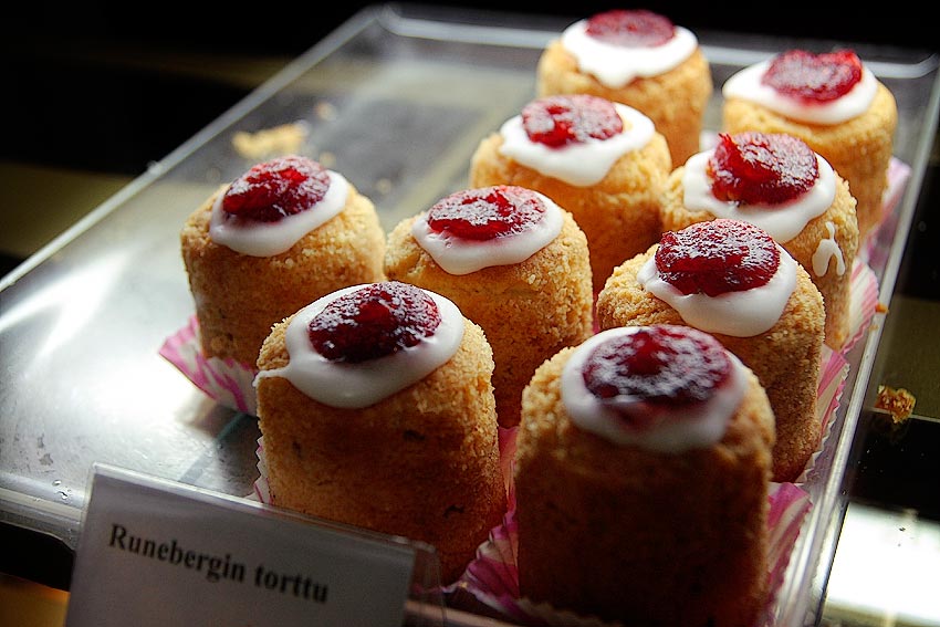 A close-up photograph of Runeberg’s torte, a moist and dense pastry with a tender crumb, topped with a smooth layer of raspberry jam and white icing. The cakes appear nutty and slightly textured, sitting on a tray. Photo credit: Tony Öhberg/Finland Today.