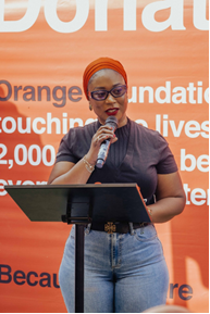 Woman in an orange hijab speaks into a microphone at a podium on an orange foundation backdrop.