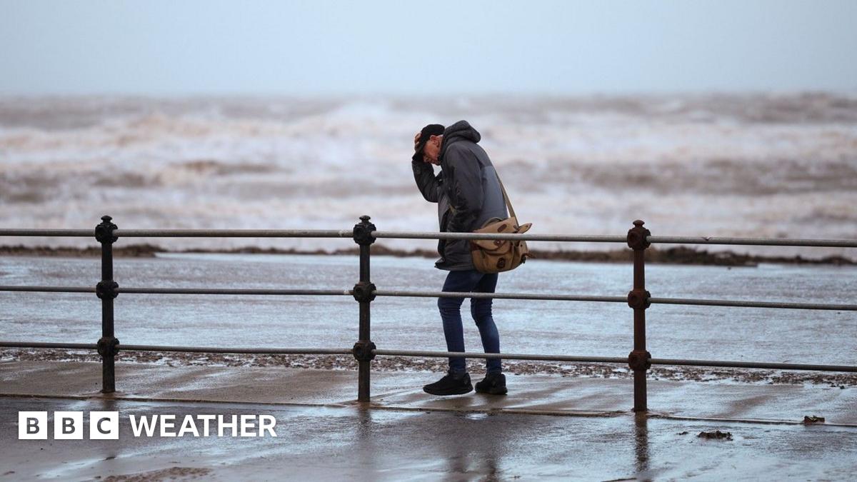 A man clutches his hat while walking along a windy seafront with rough waves in the background