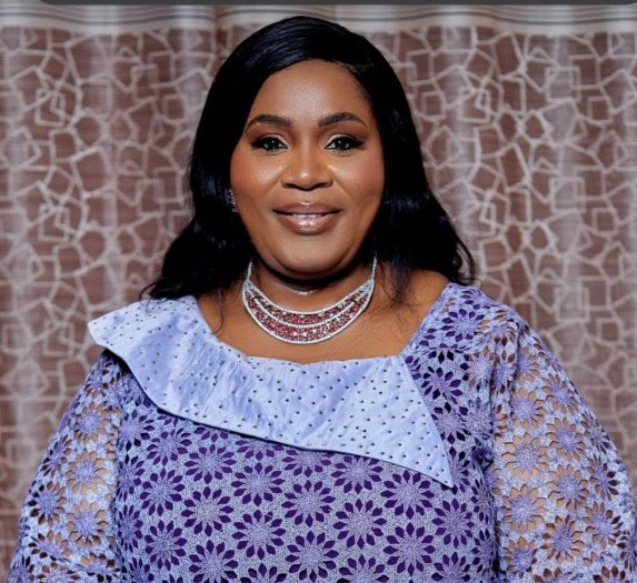 Portrait of a smiling woman in a lavender lace dress with a silver choker necklace, standing before a patterned backdrop.