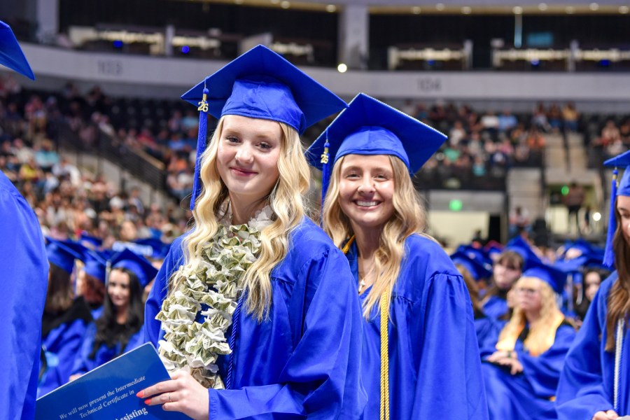 Two female students in blue graduation gowns with crowd in the background.