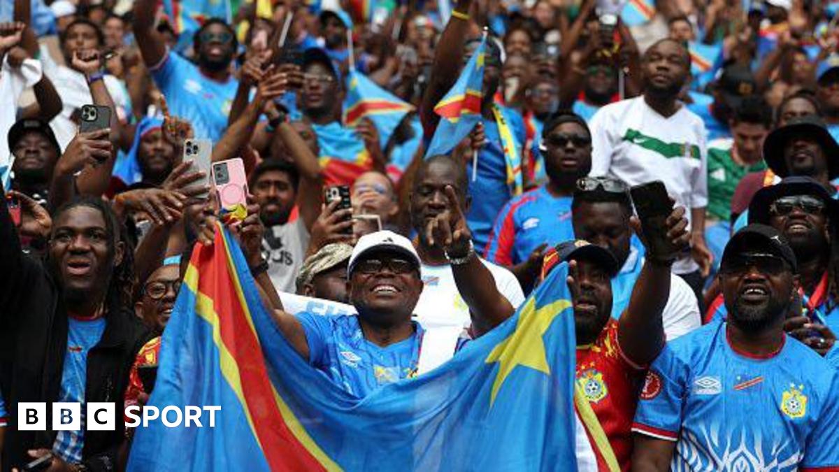 A crowd of DR Congo fans are seen in the stands cheering for their team during a 2026 Fifa World Cup play-off match against Jamaica. A fan in a white cap and blue shirt is in the foreground holding up a DR Congo flag, with many behind him holding up phones to take pictures