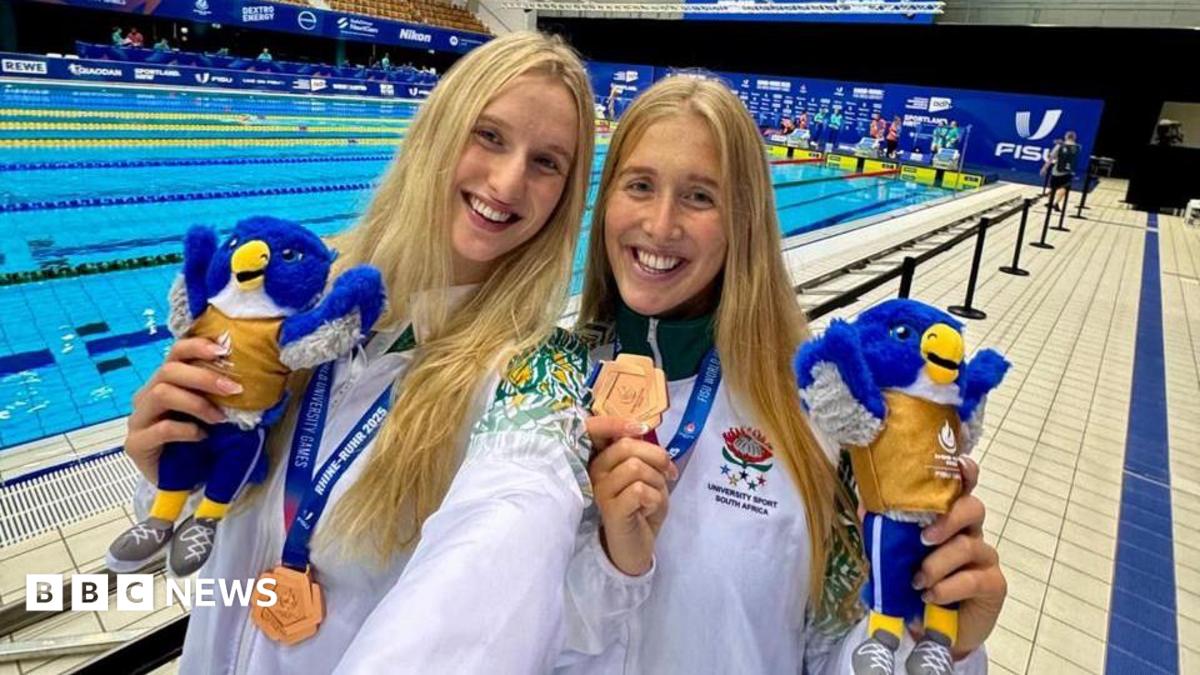 Olivia (left) and Georgia Nel stand in front of a swimming pool. They both have long blonde hair and are wearing white tracksuits, carrying toy birds and wearing gold medals.