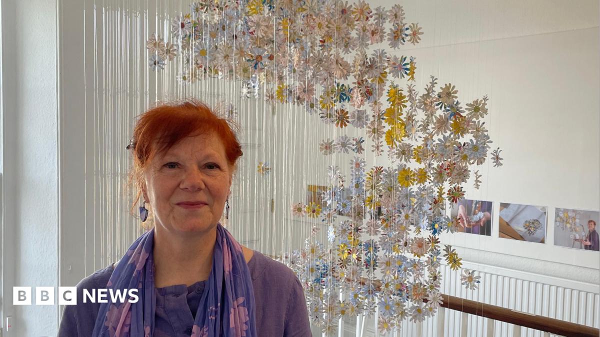 Gillian Taylor, who has red hair and is wearing purple, stands in front of her art installation. The daisies made from the letters hang on fishing wire from the ceiling of the stairwell.