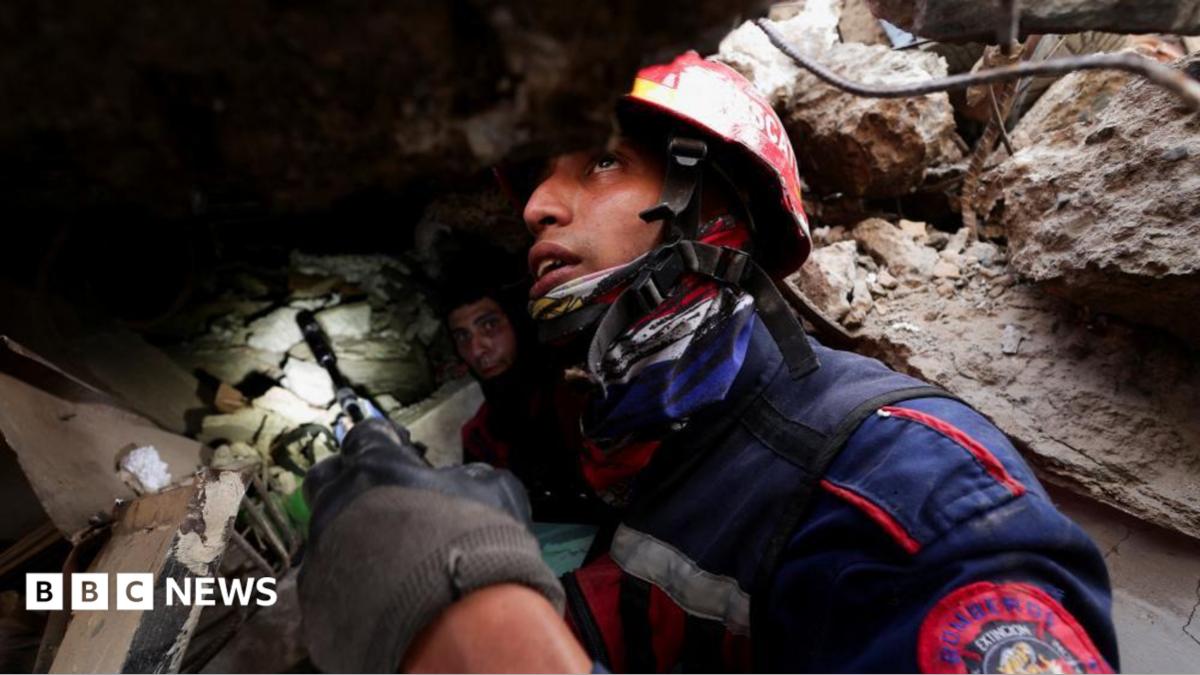 the picture shows half of the boys face - the rest is obscured by rescue workers. He's wearing a white hard hat and has dark curly hair.