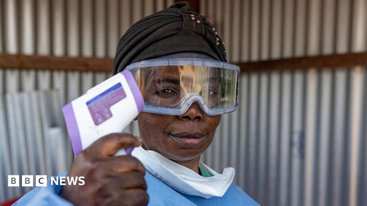 A healthworker, wearing goggles and a mask, pulled down from her mouth, holds up a thermometer