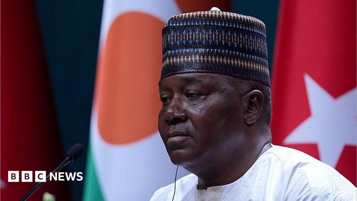 A head and shoulders image of Abdourahamane Tchiani with him looking to the left of the picture. He is wearing a traditional cap and, out of focus, the flags of Niger and Turkey can be seen behind him