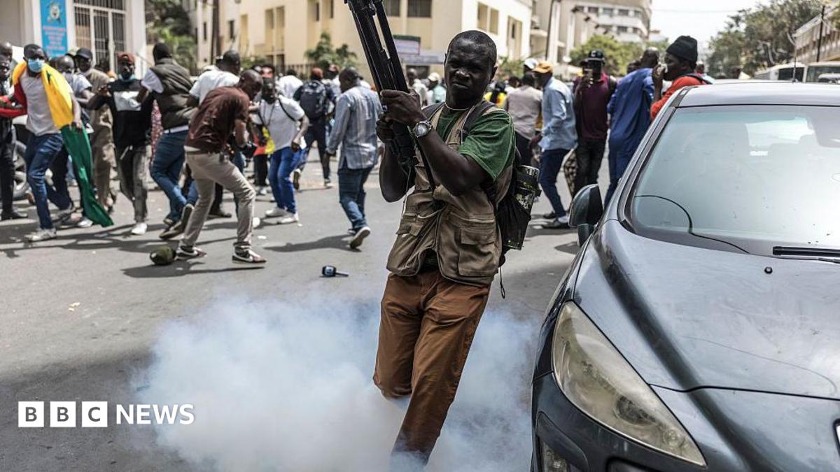 A man carrying a tripod escapes from a tear gas as smoke fills a street, while protesters scatter and gather in the background during clashes