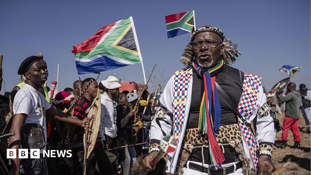 Hundreds of protesters marching outdoors, chanting and holding south african flags and signs