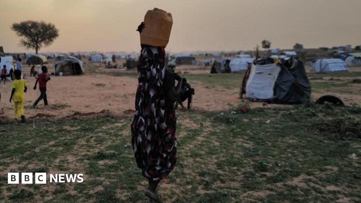 A woman, with her back to the camera and carrying a drum on her head, is walking in a camp, with children seen not far from her