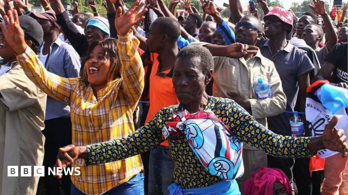 Two women in the foreground of a crowded scene hold their hands out in joy. One woman has an orange checked shirt, the other wears a Chadema party scarf.