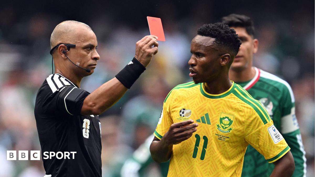 Referee Wilton Sampaio, wearing a black shirt and black sweatband on his wrist, holds a red card with his right hand in front of Themba Zwane, who looks uncomprehendingly at the official while wearing a yellow South Africa shirt with green trim and a green number 11 on the chest. Both men are visible from waist up. A Mexico player in a green shirt is half obscured in the background, with a crowd visible out of focus in the distance