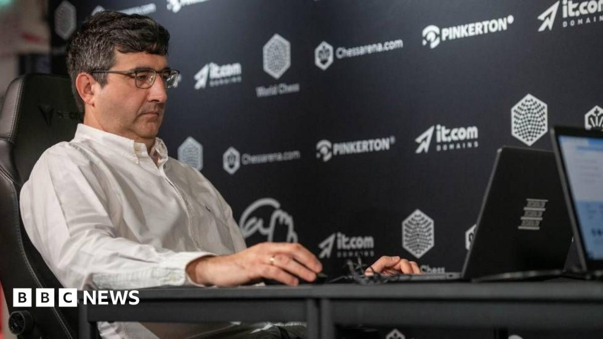 Vladimir Kramnik at a chess tournament in London in August 2024. He sits facing his opponent and is wearing a white shirt and glasses.