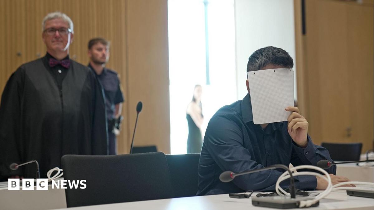 A man holds a sheet of paper over his face, wearing a black shirt in court