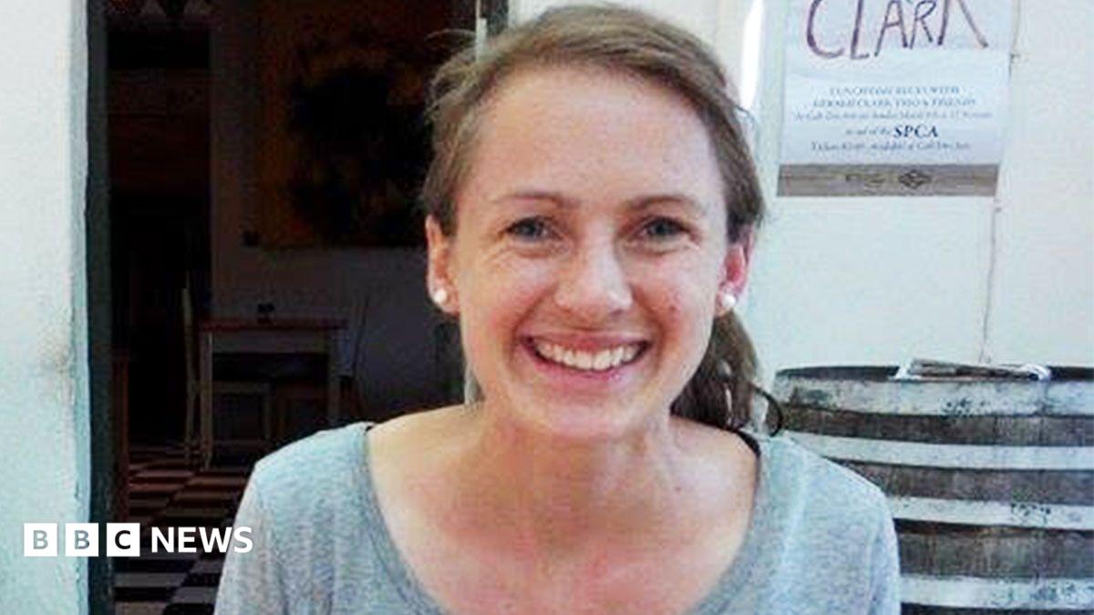 A woman with long brown hair, tied back in a ponytail, smiling at the camera. She is wearing pearl-like earrings and a scoop neck grey top.