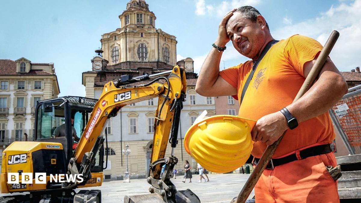 A construction worker wearing orange hi vis uniform holds a yellow construction helmet and wipes his head in front of a JCB digger next to a public square in Turin, Italy on 25 June.
