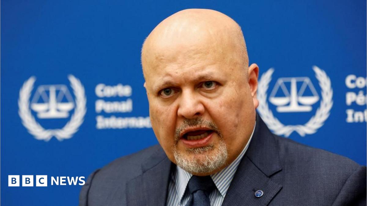 International Criminal Court Prosecutor Karim Khan speaks during an interview while sitting in front of a blue sign with blurry white writing and the symbol of the ICC - the scales of justice surrounded by a laurel wreath.