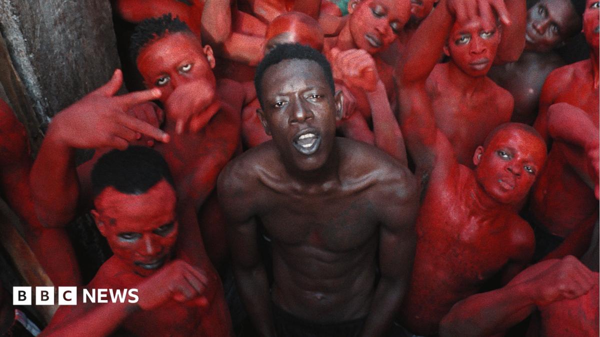 A topless Empereur stands in the centre of a group of young men, who are painted in red.