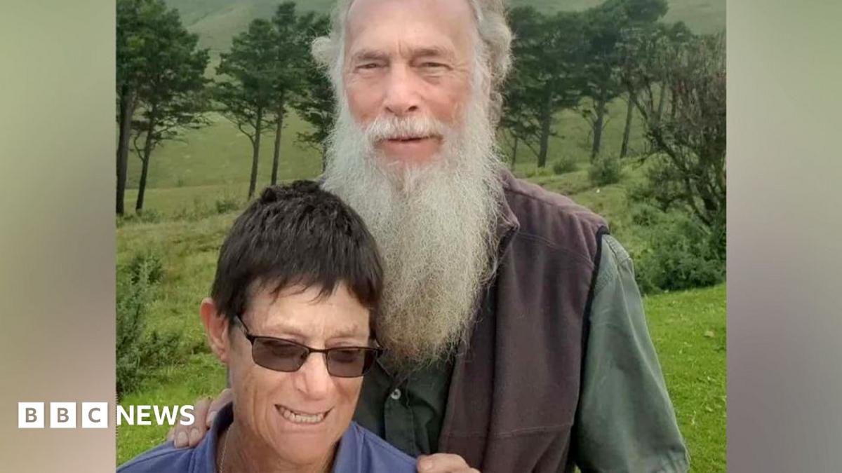 Rodney Saunders stands behind Rachel. Rodney has a long grey beard while Rachel has dark hair and is wearing sunglasses