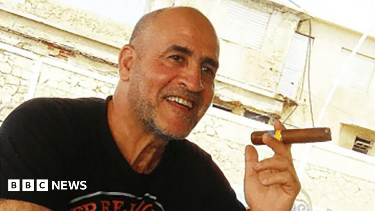 Daniel Siad, a man with a shaved head and stubble, wearing a black T-shirt, sits outside in what appears to be a sunny street, smiling with a large cigar raised in his hand.