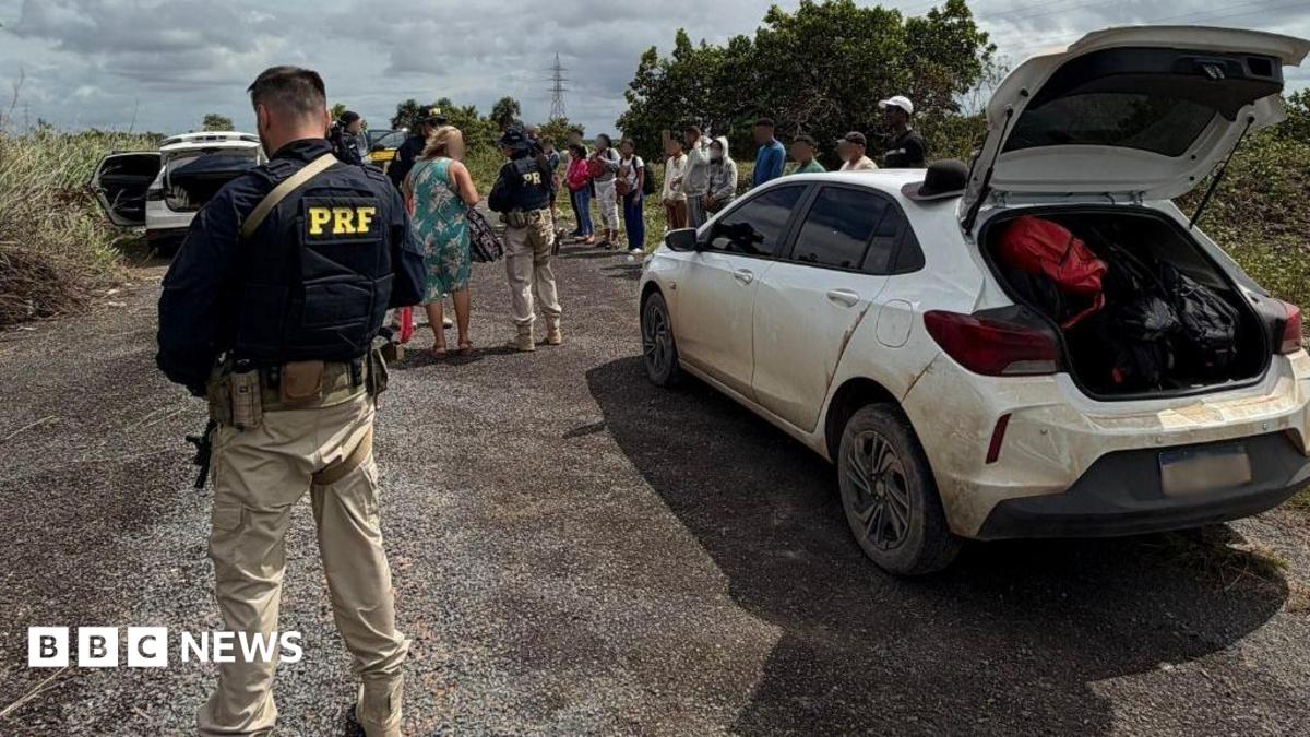 A federal highway policeman in Brazil is seen in a bullet-proof vest in the foreground, stood a few yards from a white dusty car that has its boot open. In the background, an officer can be seen talking to a woman, while a row of about a dozen people is lined up on the roadside. Another vehicle with its boot and doors open can also be seen in the background.