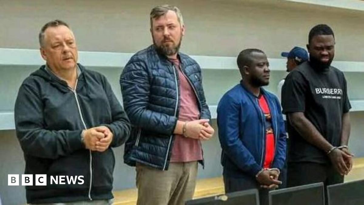 Four handcuffed men standing in front of a row of confiscated laptops. Lev Lakshtanov, Igor Ratchin, Amor Carlos Tomé and Francisco Oliveira (left to right)