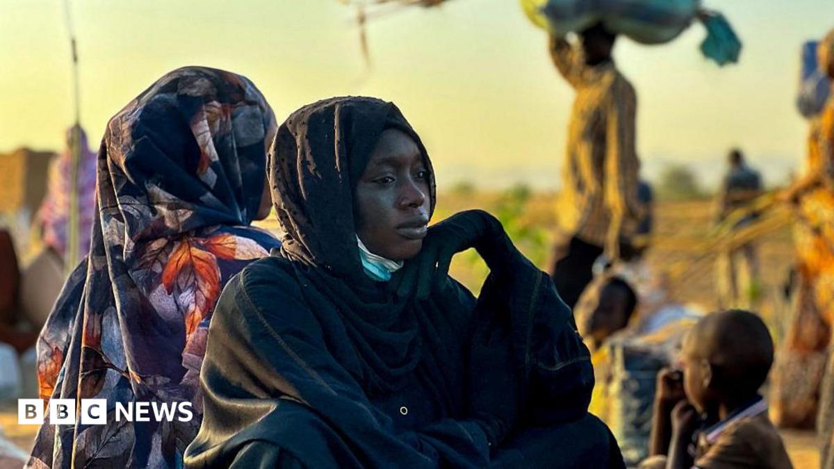 A woman in a black headscarf sits thoughtfully as people and children gather in the background.