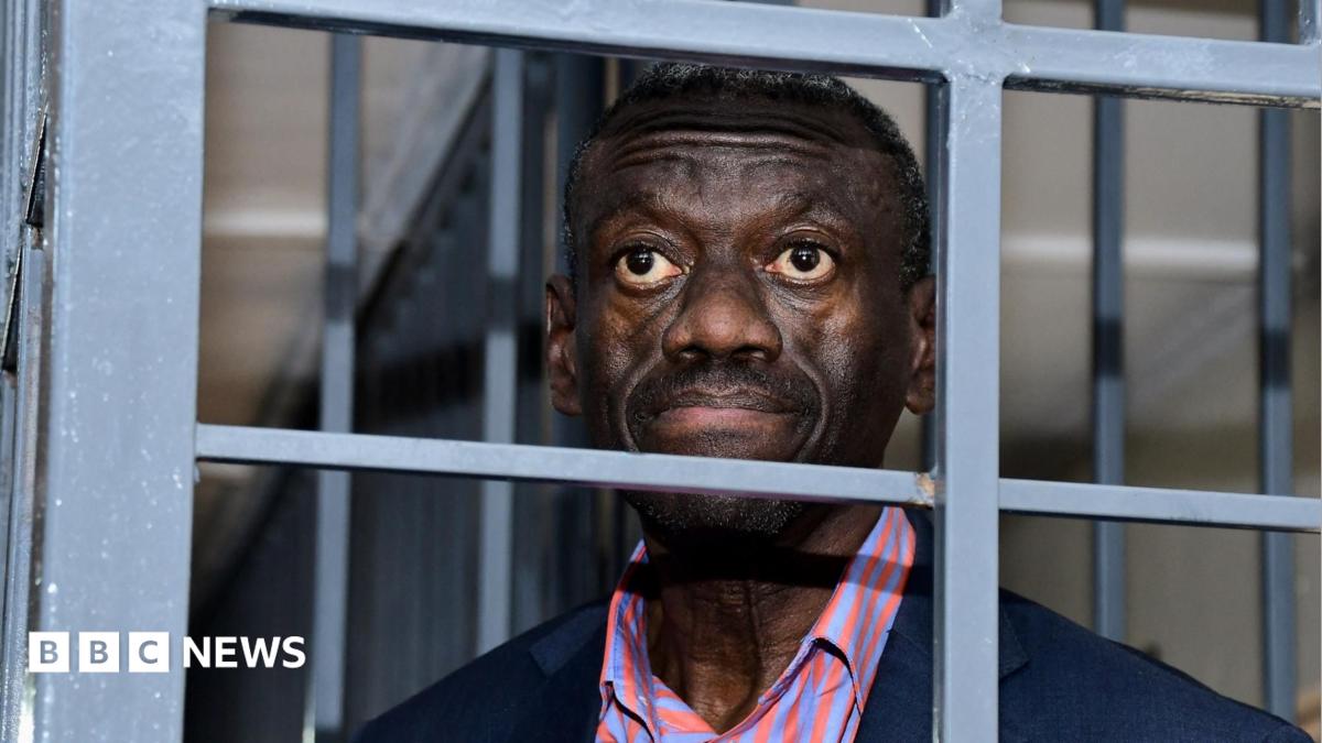 Kizza Besigye and a salmon pink and purple stripped shirt and a navy jacket seen behind bars