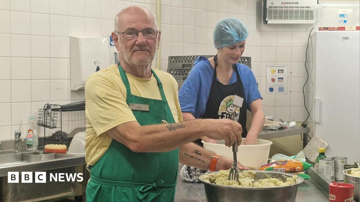 Ian White with glasses, a yellow t-shirt and green apron, preparing mashed potatoes in a large bowl while a woman with a blue hairnet, blue t-shirt and blue apron works nearby in a kitchen.
