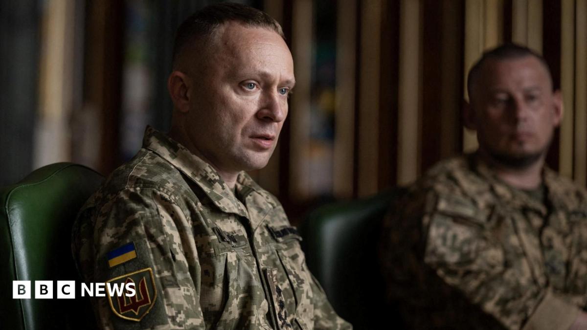 A man sits wearing a military uniform while another uniformed man looks on in the background