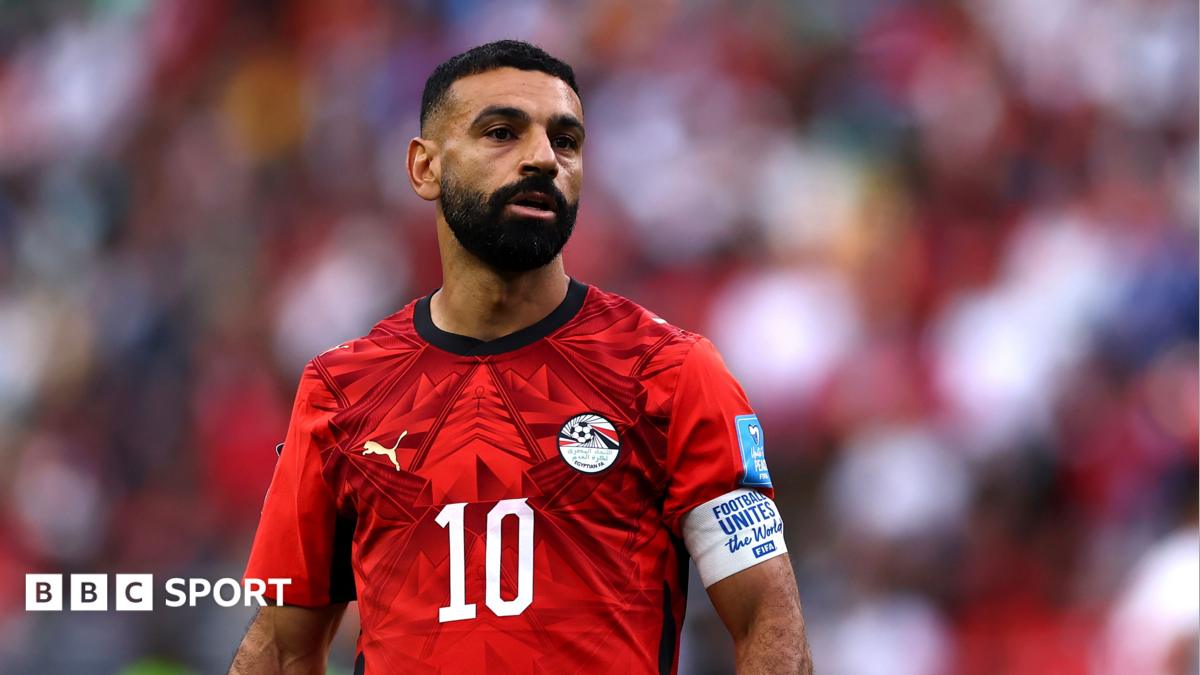 Mohamed Salah, visible from waist up, in training with the Egyptian national team at the World Cup in the USA. Salah wears a red training top with white lines running from armpit to neck on both sides, a white sponsors logo and the crest of the Egyptian FA. The entirety of the background is taken up by a white net with a white goalpost visible to the left of Salah