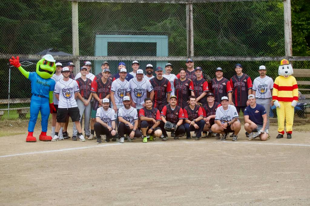 The Stilly Valley Little League Dads and Snohomish County Fire District 21 Stations 49 & 50 pose with Everett AquaSox mascot Webbly (far left) and Sparky the Fire Dog (far right) ahead of their fundraising softball matchup at the Arlington Heights Baseball Field on Aug. 21, 2026. (Joe Pohoryles / The Herald)