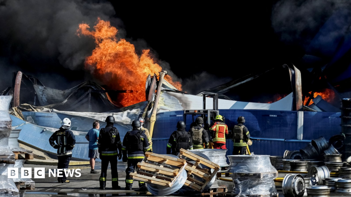 Wide shot showing Ukrainian firefighters in black and yellow uniforms observing flames rising from the debris of what appears to be a warehouse following a drone strike.
