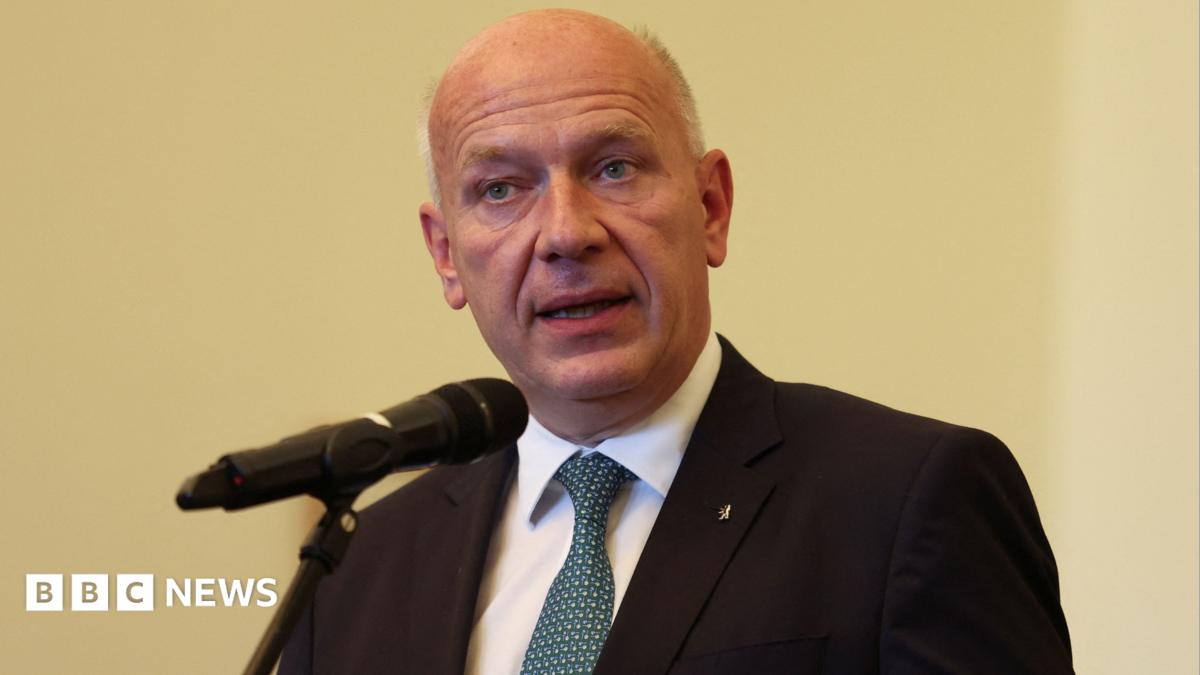 Berlin Mayor Kai Wegner speaks into a microphone on a pale yellow background wearing a dark suit and blue tie.