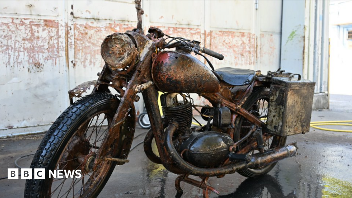 A rusted but largely well-preserved World War Two-era Wehrmacht motorcycle pictured parked in a building resembling a warehouse.