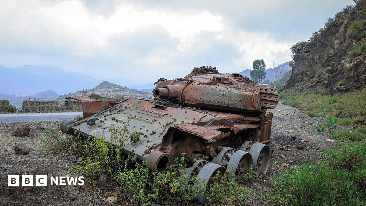 A rusted, wrecked tank lies on the side of a rural road. Mountains are visible in the background.