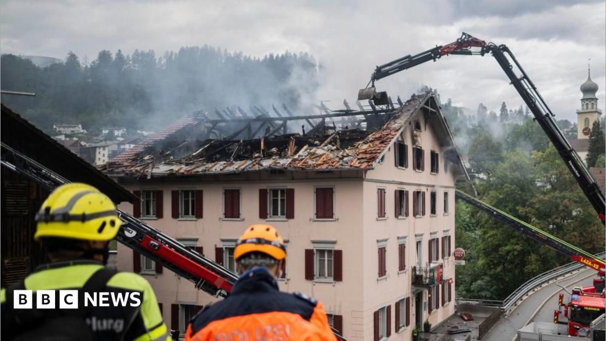Large flames come out of the windows in the top floor of a building at night.