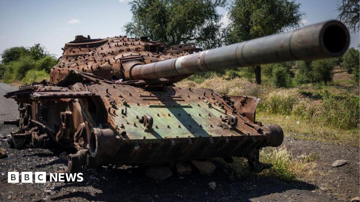 A rusted, wrecked tank lies on the side of a rural road. Mountains are visible in the background.