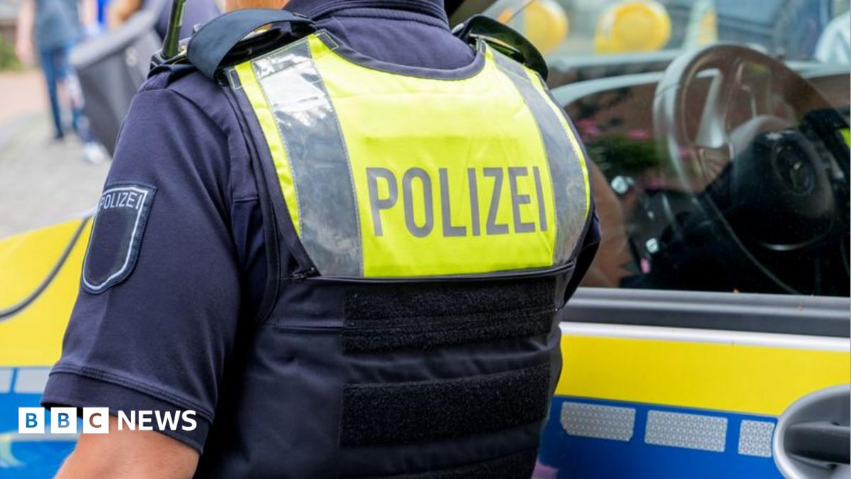 A German police officer standing next to a police vehicle. The officer wears an operational vest that reads "polizei".