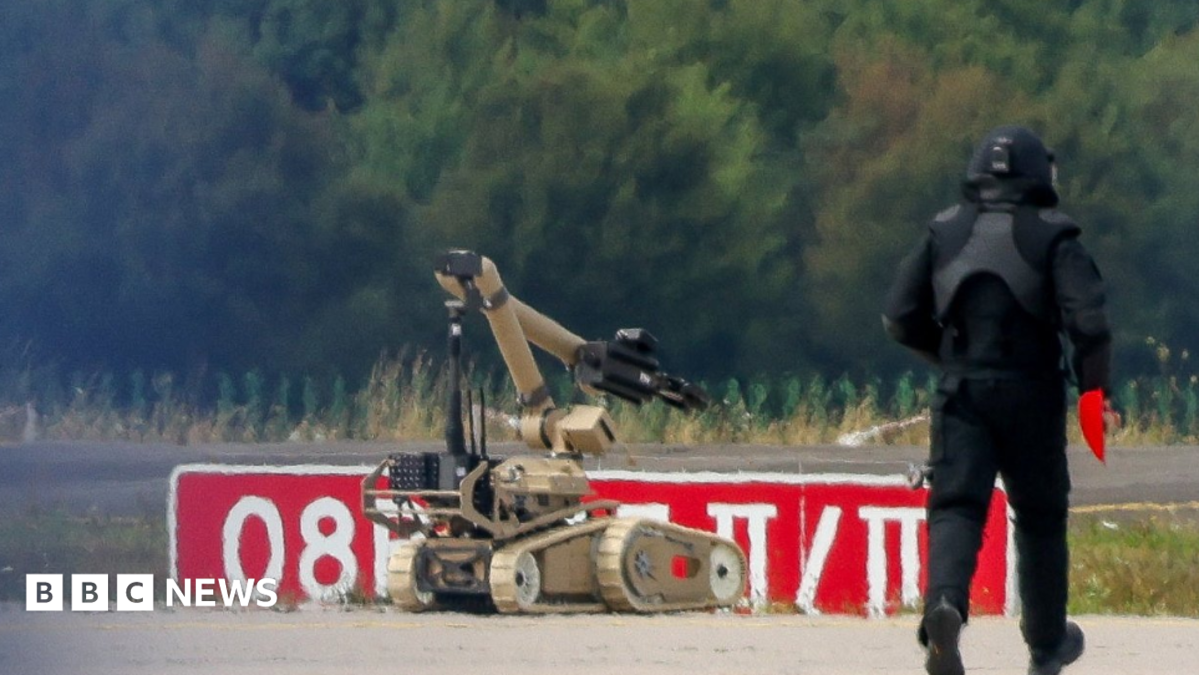A police officer in dark protective clothing walks next to an explosive-disarming robot at Leipzig/Halle Airport on 5 August 2026