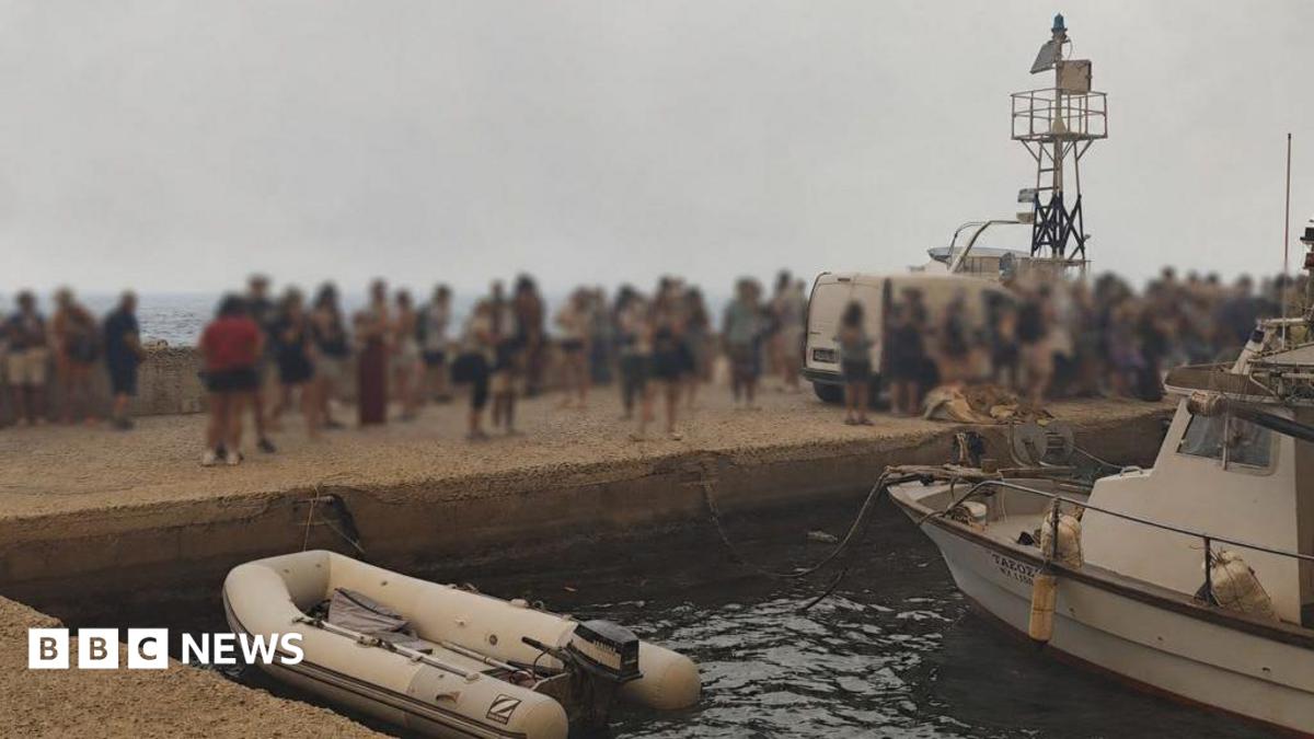 Blurred-out people stand on a marina pier while waiting to be evacuated in Agios Vasileios in Greece.