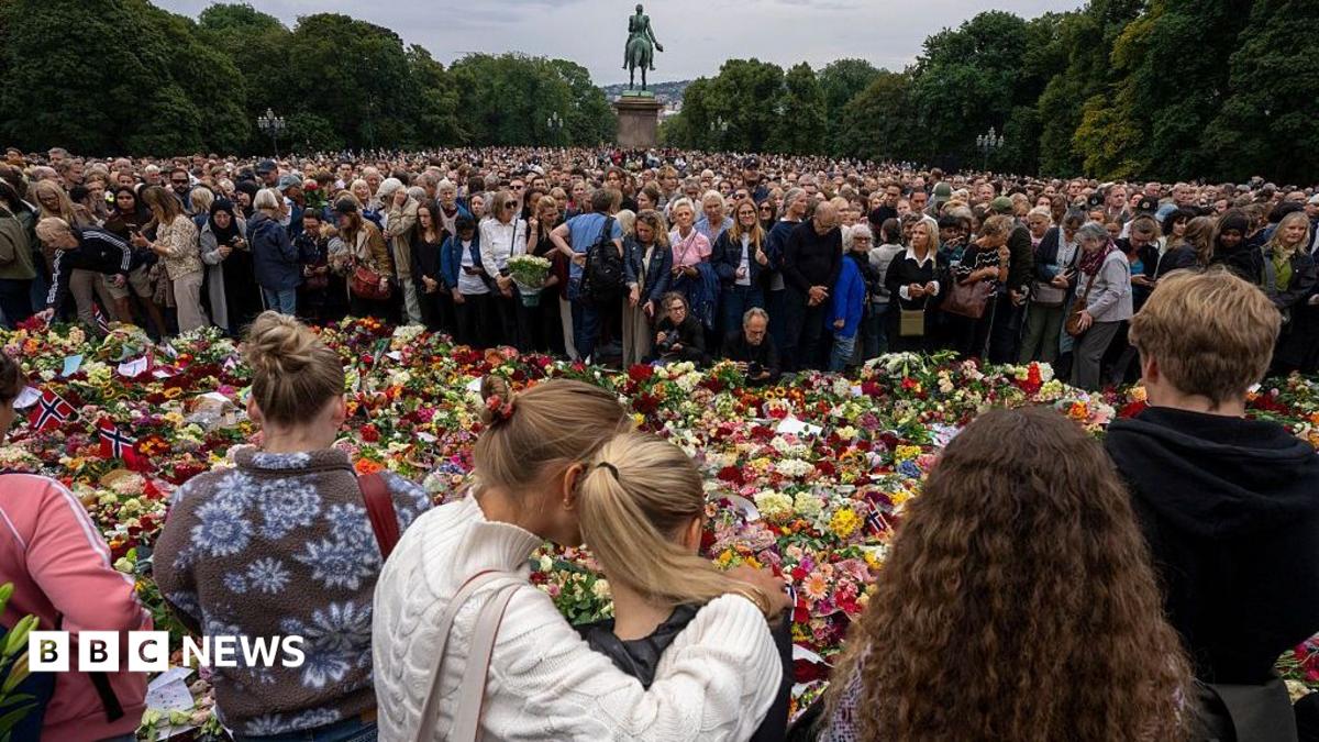 People stand in a crowd, two women hugging each other, against a line of flowers and flags on the ground, a statue behind them at the royal palace in Oslo.