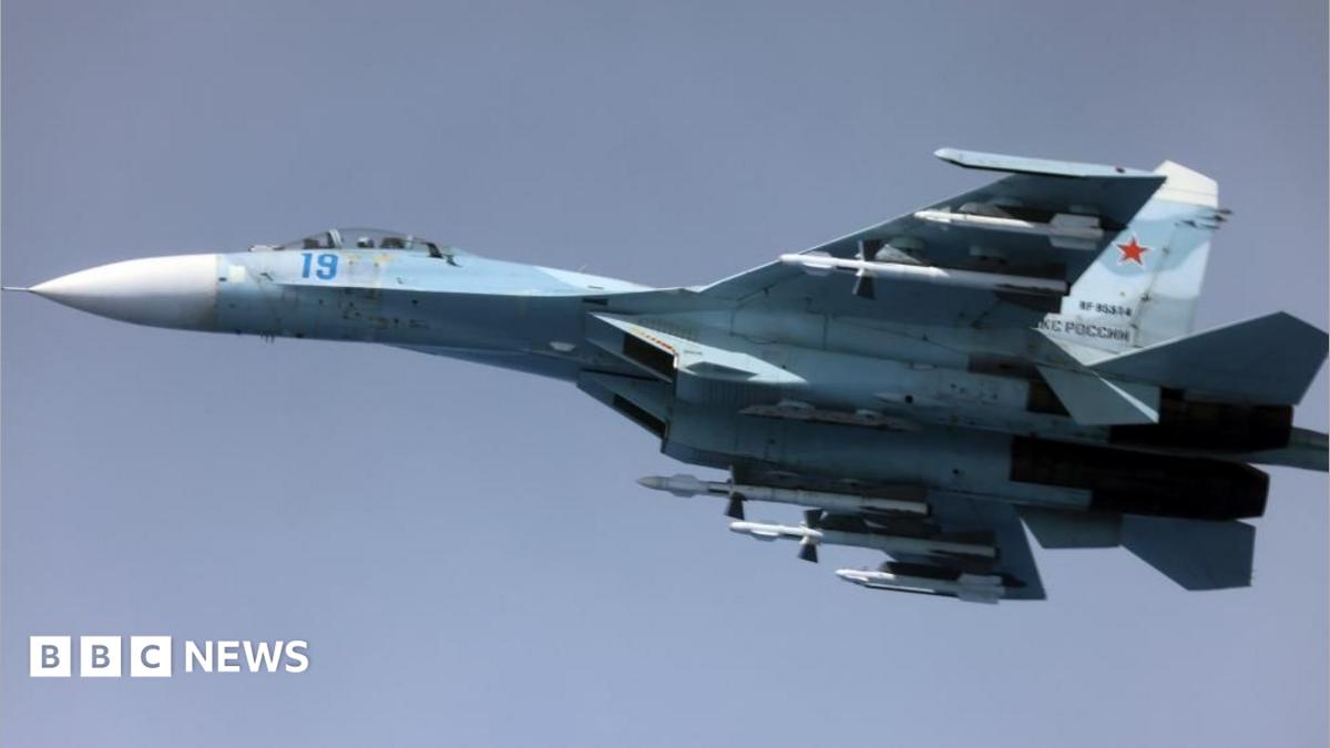 Image of a Russian Su-27, a whit and blue coloured fighter jet, airborne against a clear blue sky.